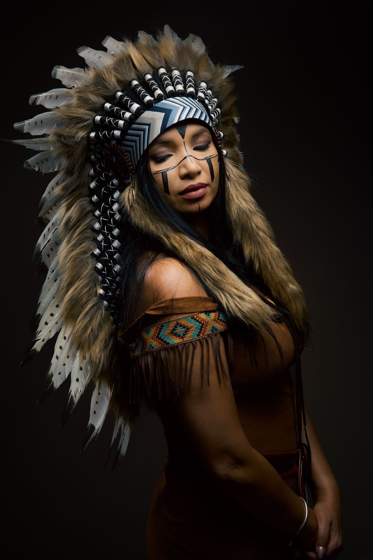 Traditional Native American headdress and fringed dress worn in dramatic studio portrait against dark background.