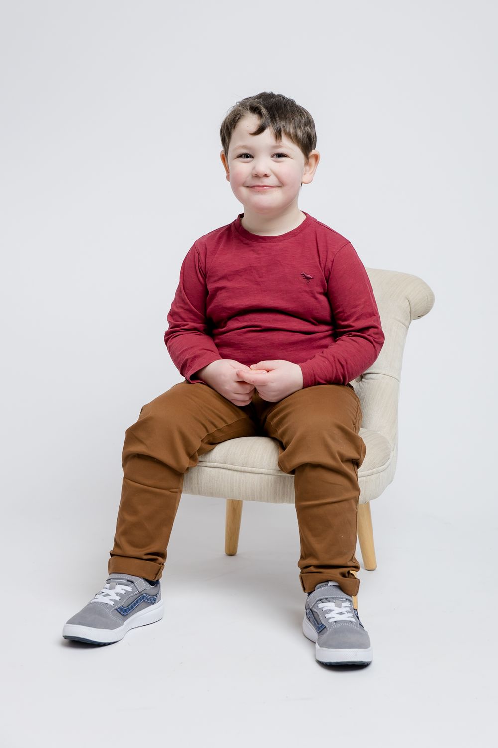 Young child in red sweater and brown pants sits smiling on beige chair against white background.