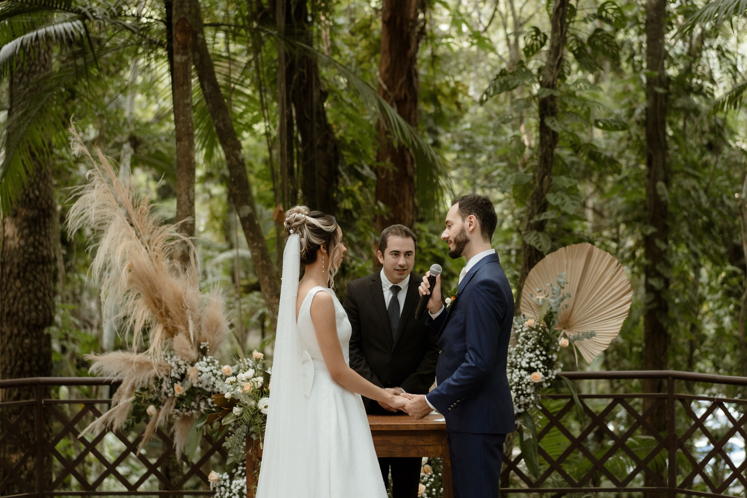 Outdoor forest wedding ceremony featuring pampas grass decor and decorative fans at a rustic wooden arbor.