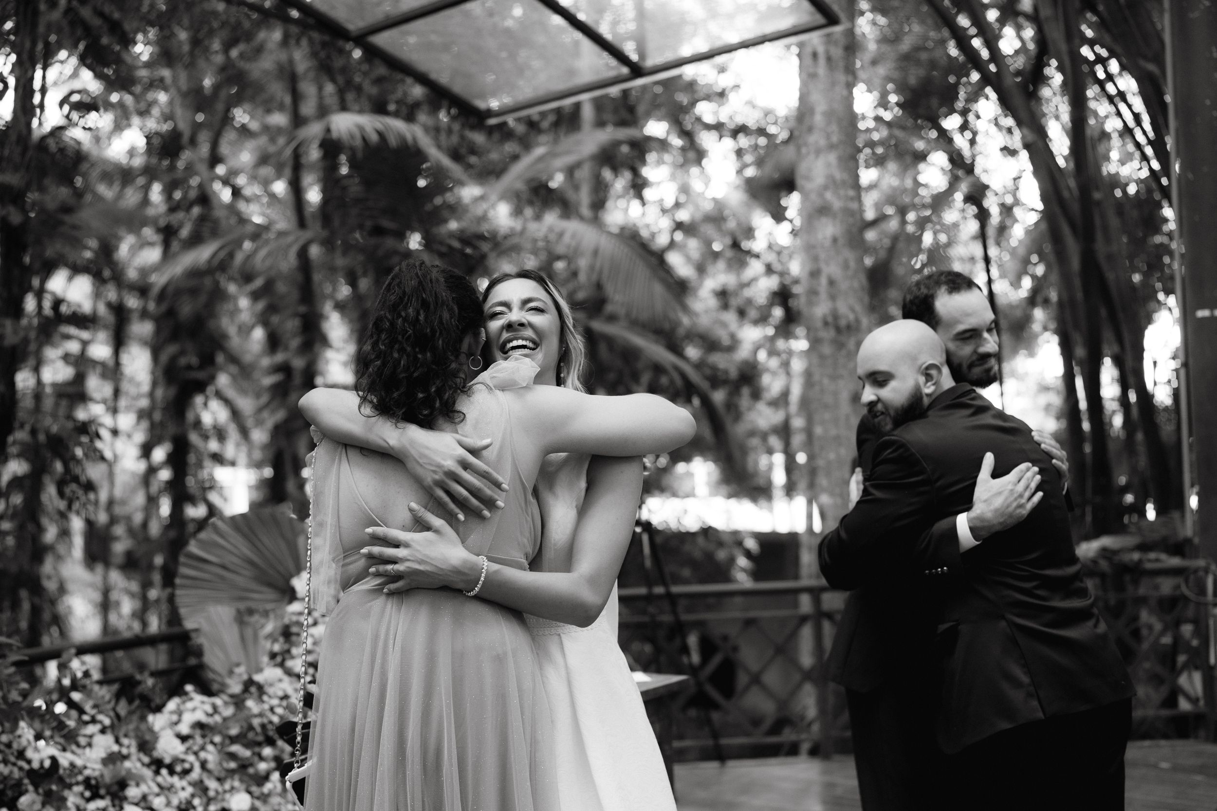 Black and white candid photo of emotional embraces during wedding celebration in forest setting.