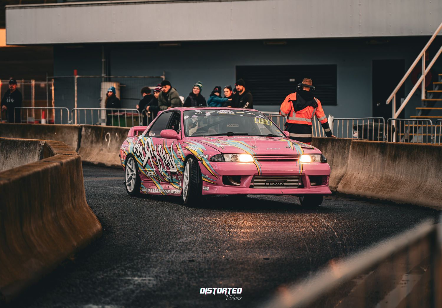 Pink modified drift car racing on wet track with concrete barriers and spectators watching in background.