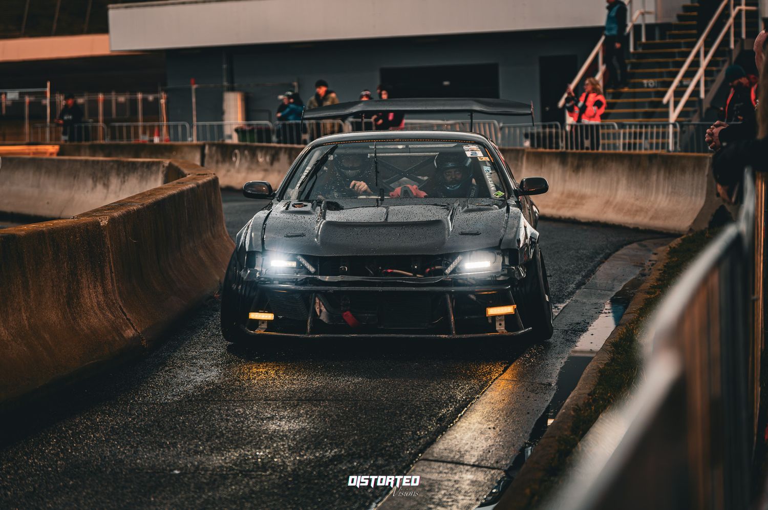Modified black sports car with custom bodywork parked on wet pavement at night with amber lighting in the background.