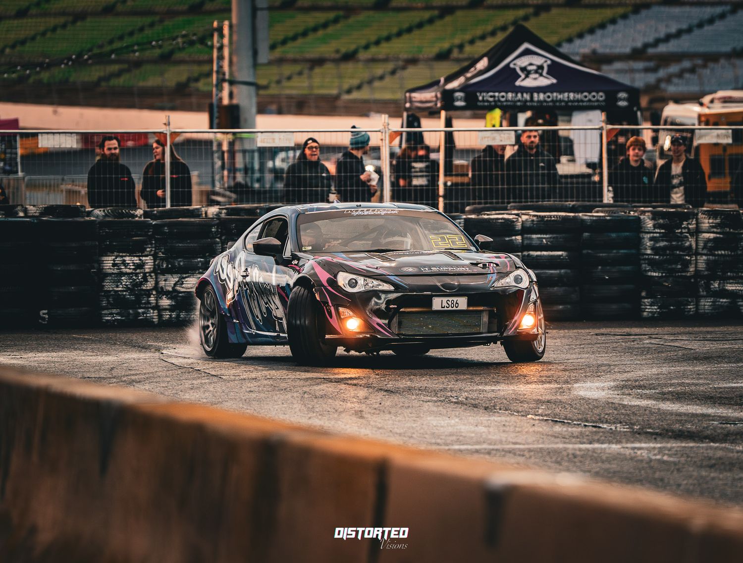 Black drift car slides sideways on wet pavement during a nighttime motorsport competition.