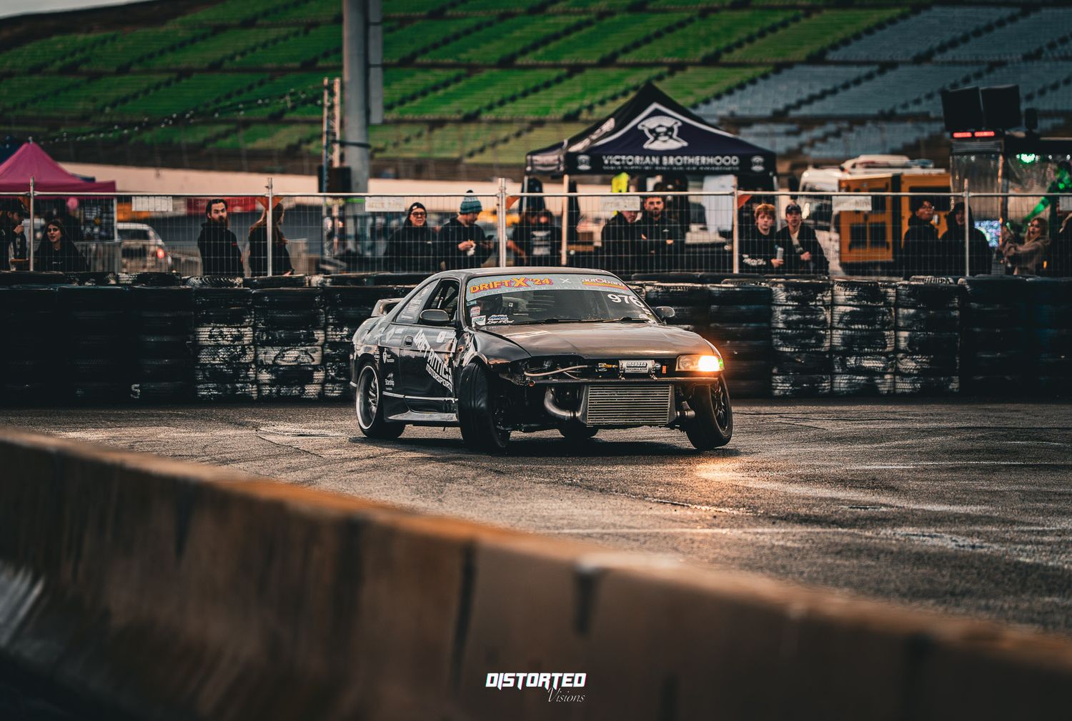 Modified black Nissan Skyline drifting at night during a motorsport event with spectators and tents in background.