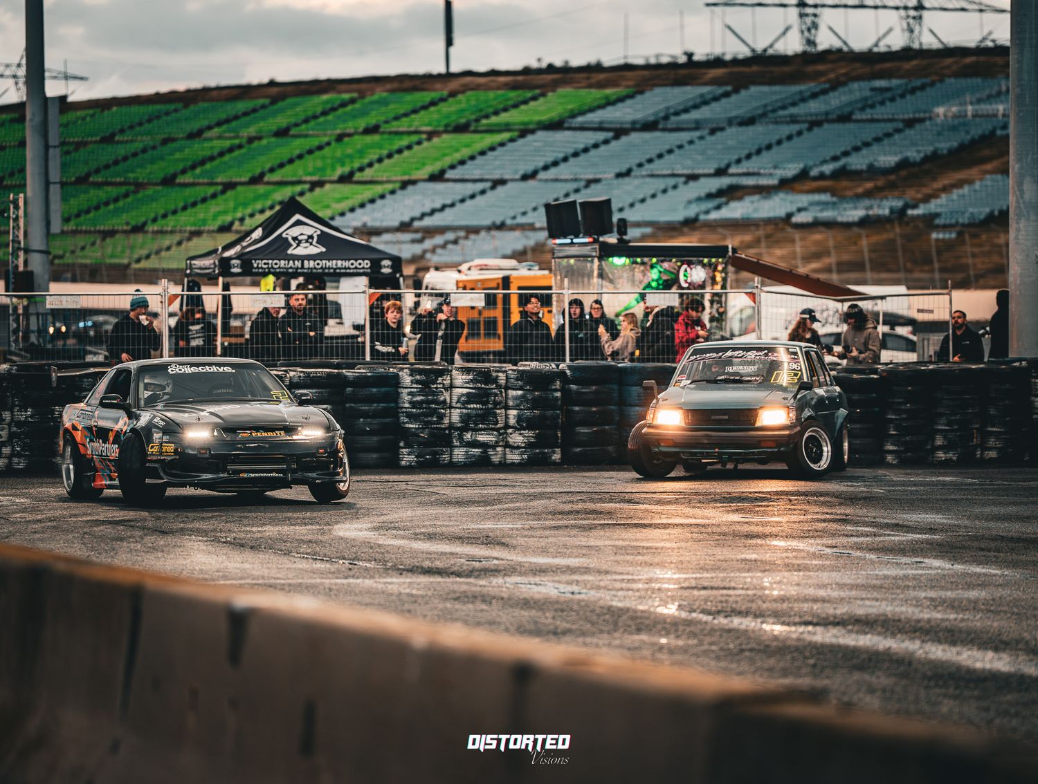 Cars with illuminated headlights gather in a dimly lit parking lot during dusk hours.