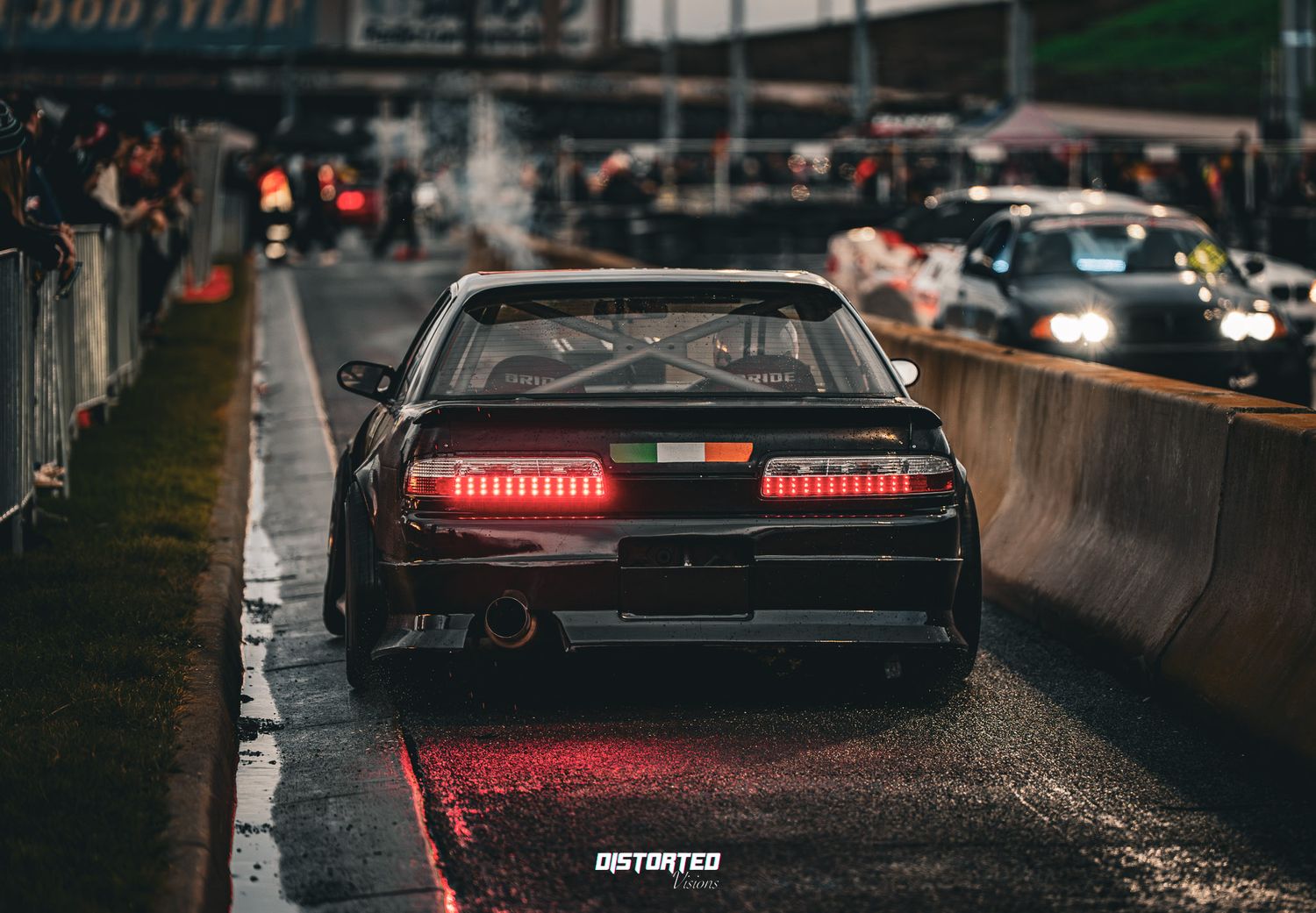 Rain illuminates red taillights on a dark city street with multiple parked cars during a gloomy evening.