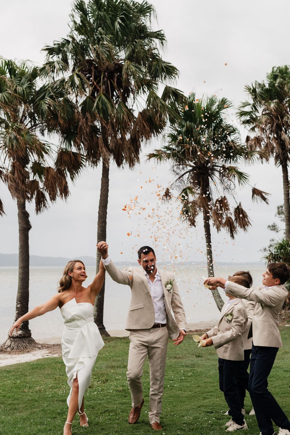 Bride and groom celebrate wedding on tropical beach lawn with palm trees, confetti, and two children in matching suits.