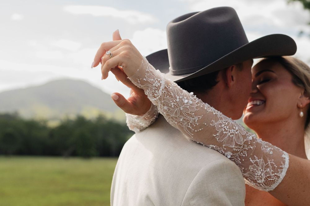 A bride in a lace wedding dress embraces a groom wearing a cowboy hat in a scenic mountain field.