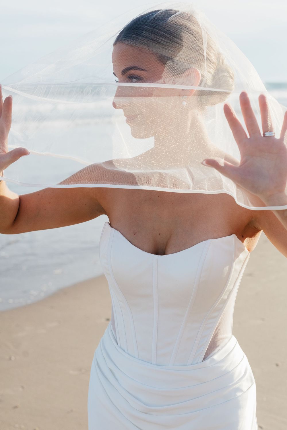 Bride in white strapless gown holds veil open on beach, wearing engagement ring, with ocean waves in background.