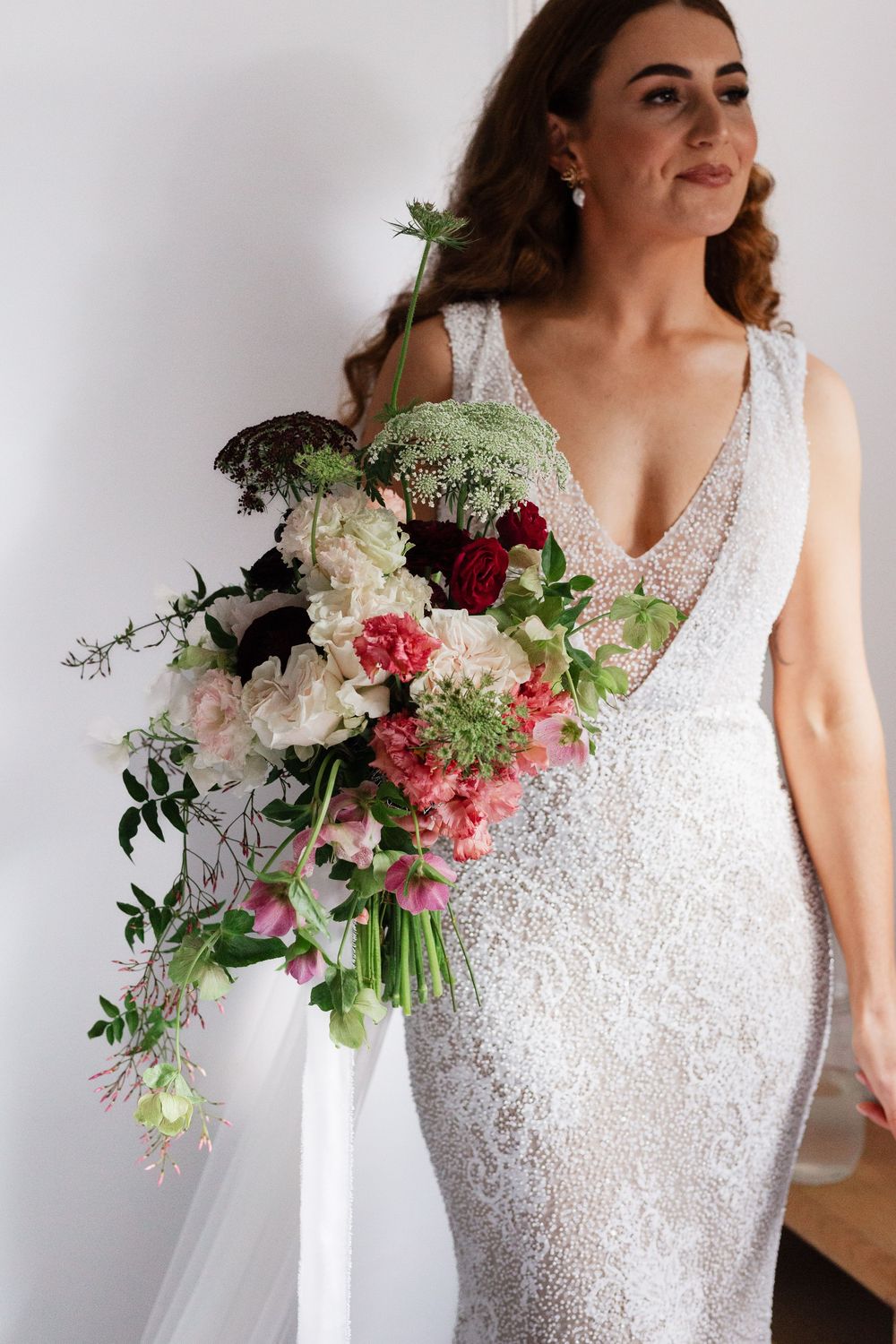 Elegant lace wedding dress with cascading pink and white floral bouquet against white wall.