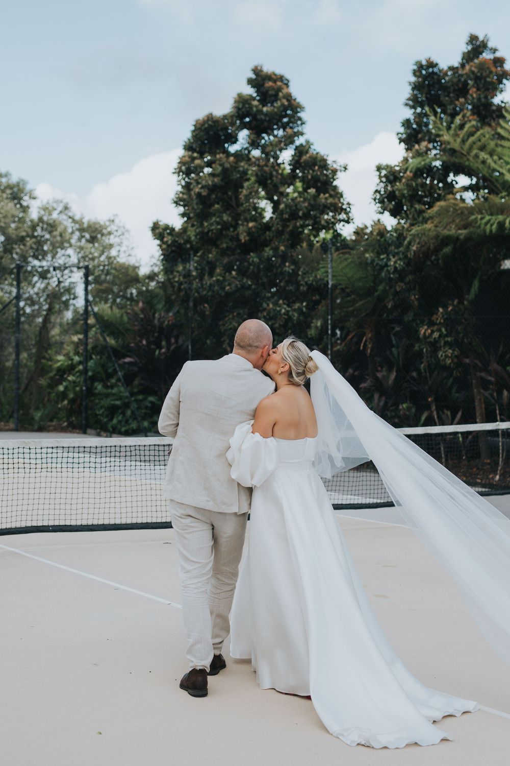 Bride and groom sharing a romantic kiss on a tennis court, her veil flowing in the breeze, surrounded by lush greenery.