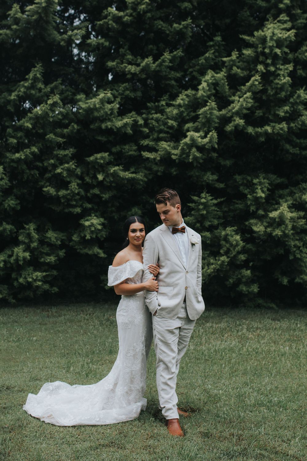 A couple poses together on grass with evergreen trees in the background.