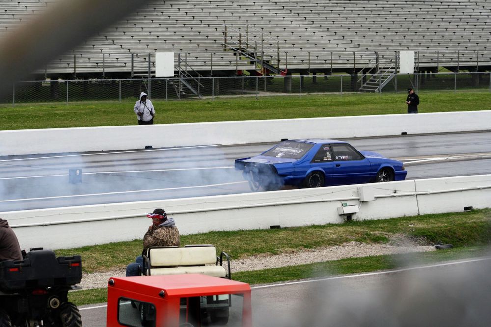 Blue race car performing a burnout on a wet drag strip surface.