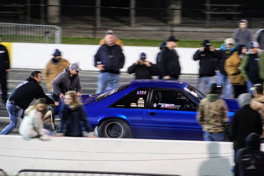 Modified blue sports cars line up at a racing event with spectators watching.
