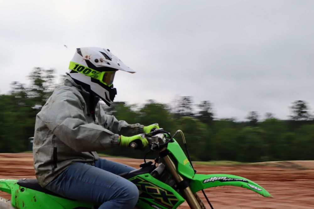 Dirt bike rider in gray jacket and neon helmet rides green motorcycle on muddy track.