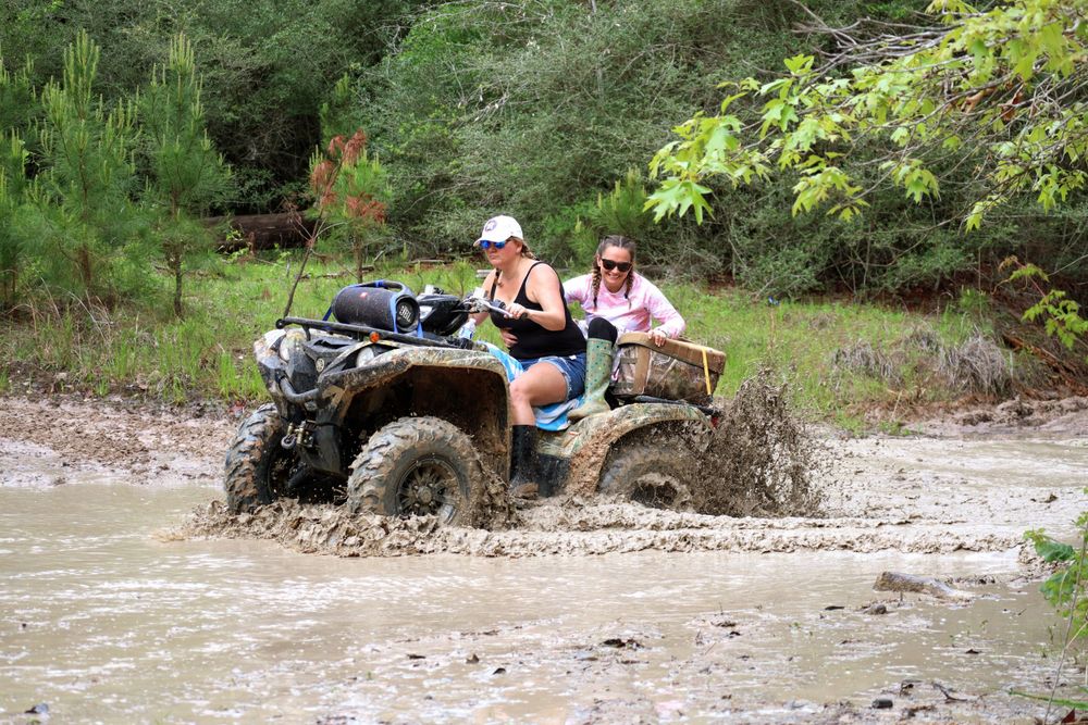 ATVs traverse a challenging muddy water crossing in a wooded setting.