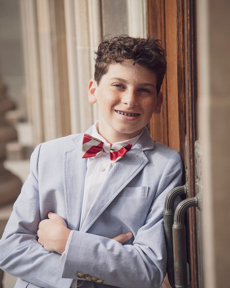 Smiling boy in light blue suit and red striped bow tie, arms crossed, leaning against wooden door.