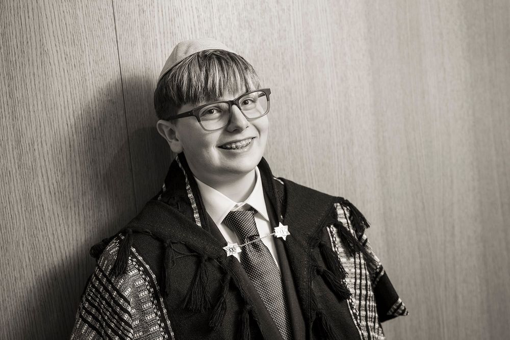 Black and white portrait of a smiling young person wearing glasses and academic graduation robes with a floral pin.