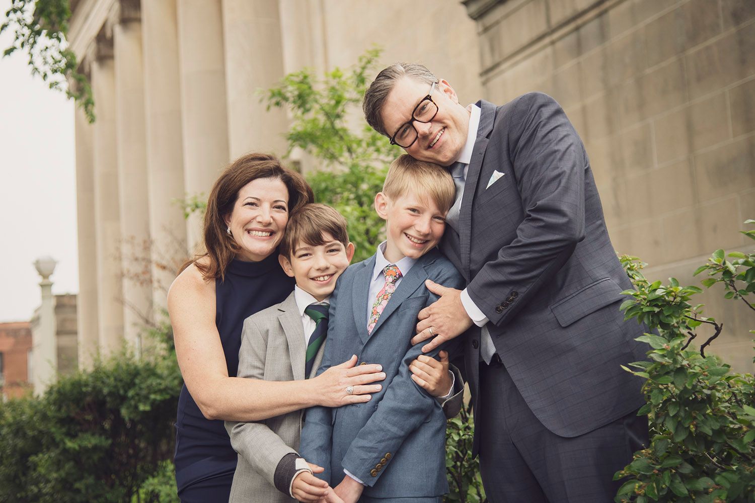 A smiling family of four in formal attire poses outdoors near stone columns and greenery.