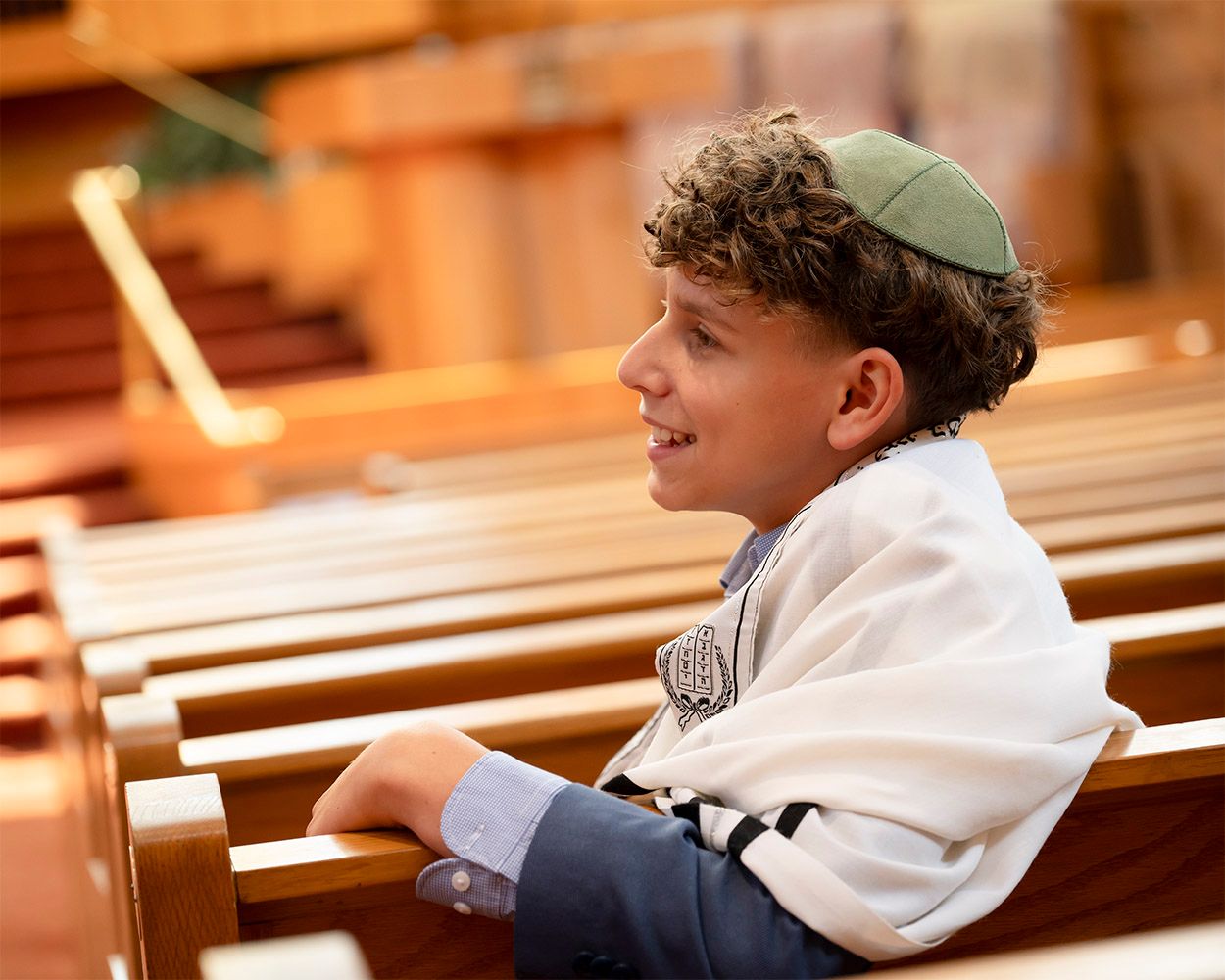 Smiling boy in green kippah and tallit sits on synagogue pew during Bar Mitzvah ceremony.