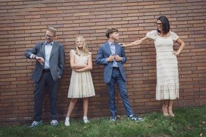 Four people in formal attire pose against a brick wall, smiling and interacting playfully outdoors.