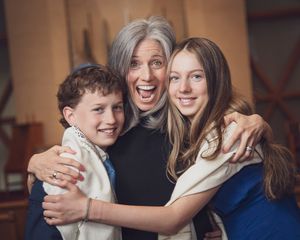 Joyful grandmother hugging two smiling grandchildren, boy and teenage girl, in a warm indoor setting.