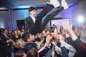 A crowd celebrates as they lift someone wearing a suit during a lively party event.