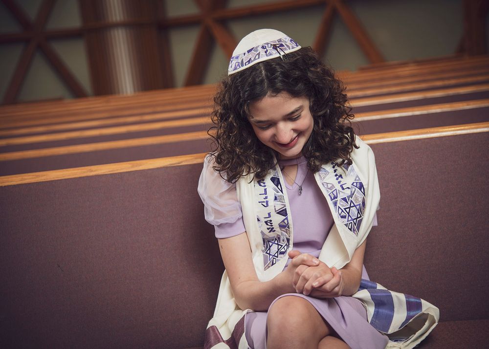 Woman in purple dress wearing kippah and tallit smiles while seated in a synagogue pew.