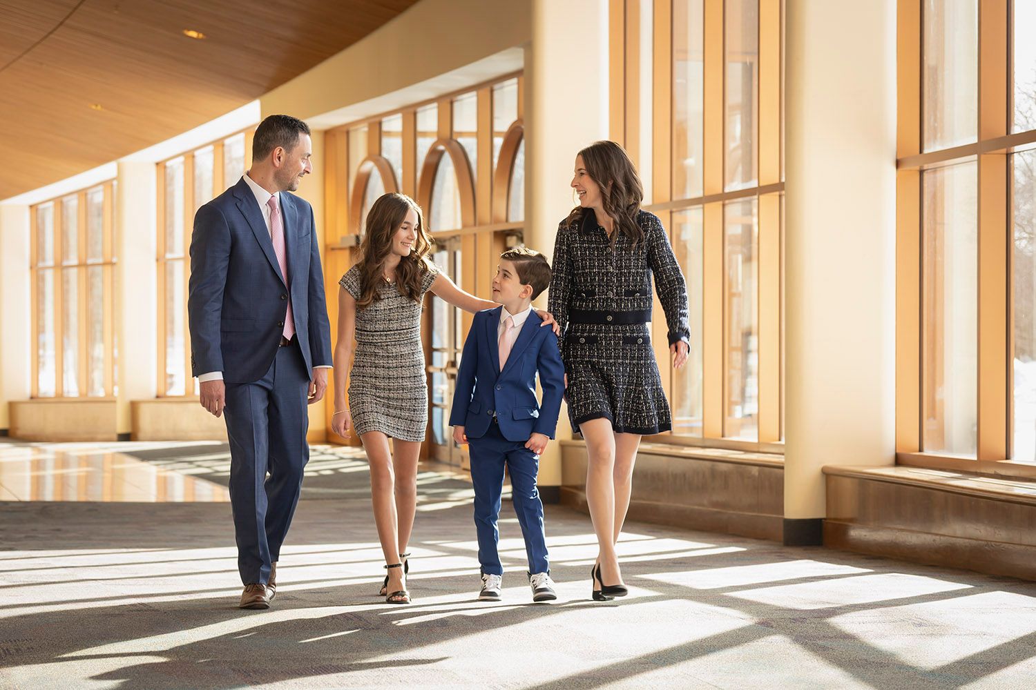 Elegantly dressed family of four walking together through a sunlit modern lobby corridor.