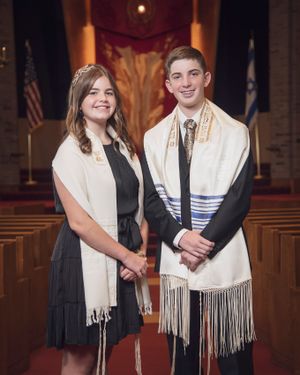 Two celebrants pose in traditional Jewish prayer shawls inside a synagogue sanctuary.
