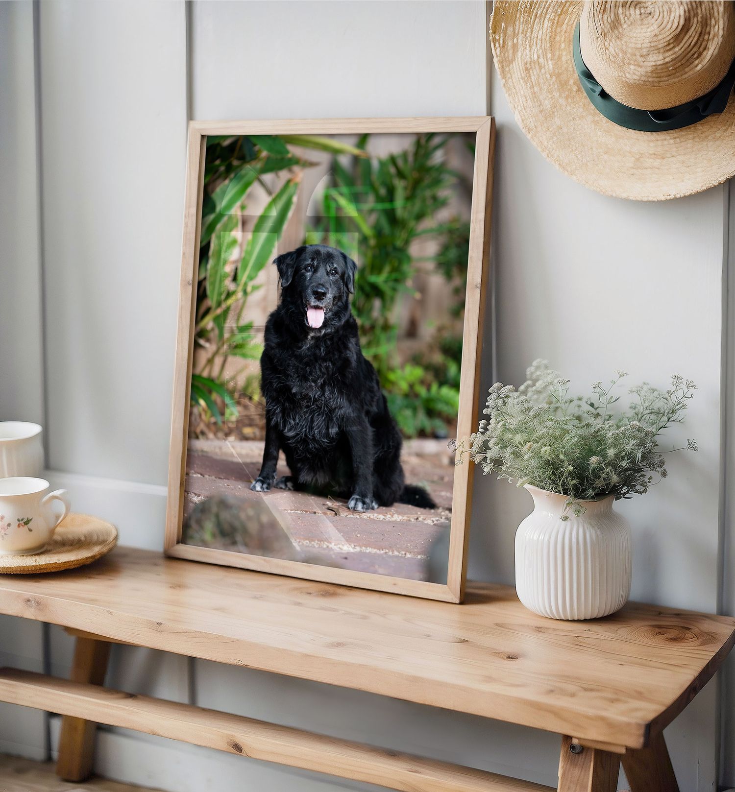 A framed photograph of a black dog sits on a wooden table with a straw hat and small vase of dried flowers.