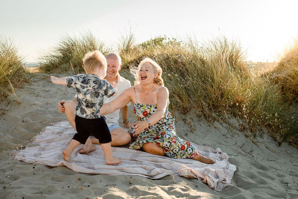 On a beach on Hayling Island, Hampshire, a couple sits in the sand at sunset, arms open as their grandson runs to them.
