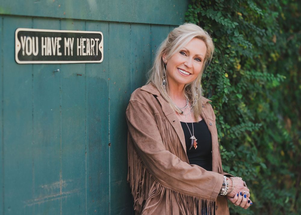 Blonde woman in fringe jacket leaning against green wall with 'You Have My Heart' sign surrounded by ivy.