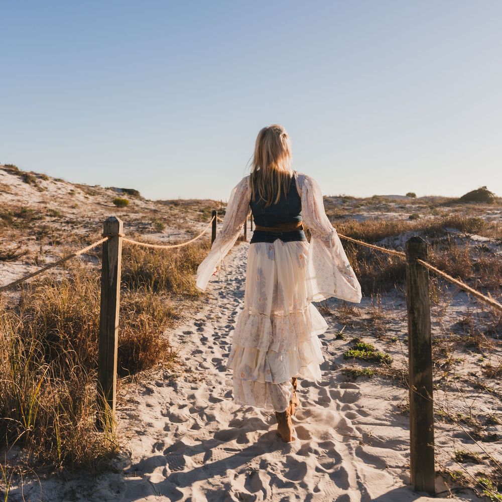 Woman in white lace skirt walks sandy beach path between rope fences toward ocean dunes on sunny day.