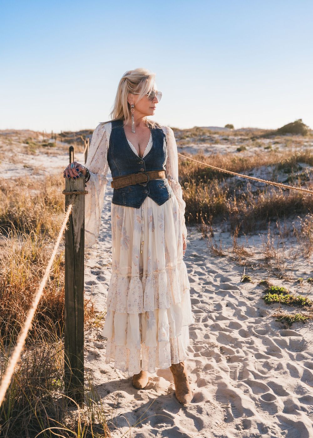 Woman in bohemian lace dress and denim vest stands on sandy beach path, posing near wooden post.