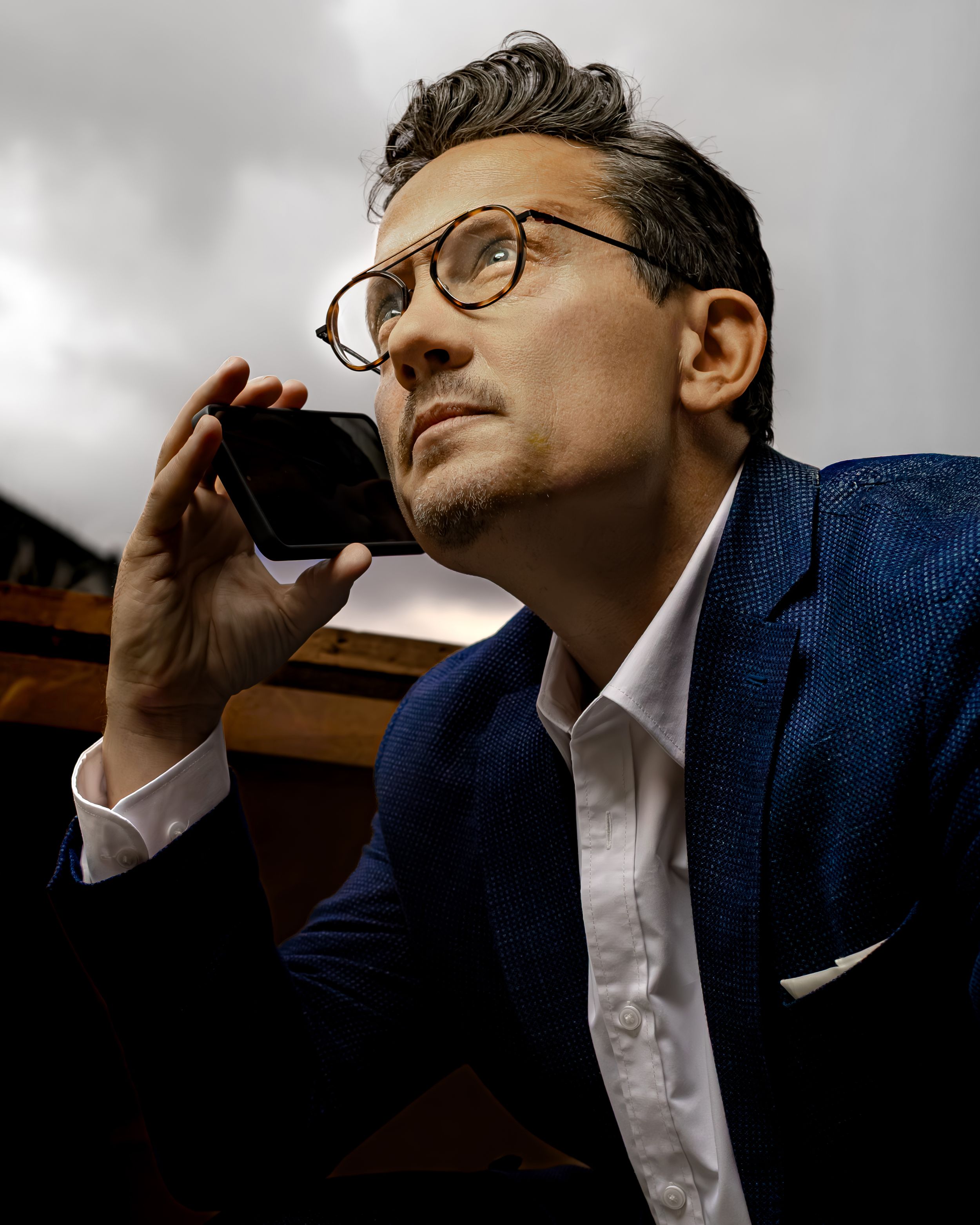 Man in blue blazer and round glasses holds phone near face, gazing upward thoughtfully against dramatic sky.