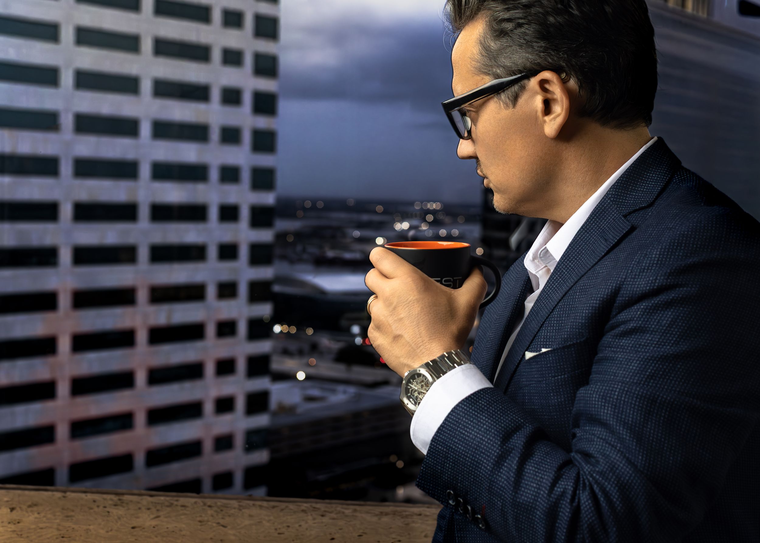 A sophisticated man in a navy suit sips a cocktail on a city rooftop balcony at dusk, wearing glasses and a luxury watch.