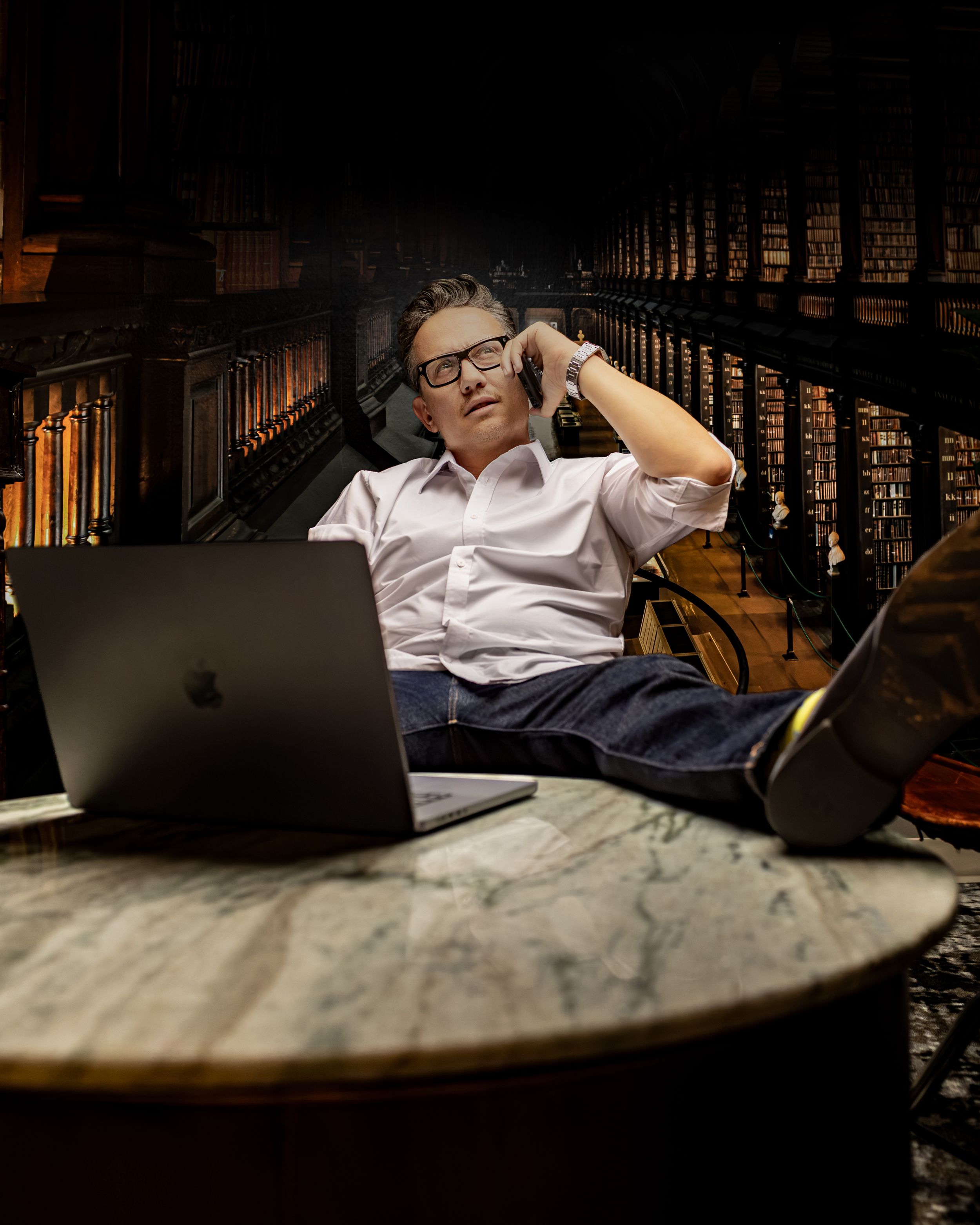 Man with headphones sits thoughtfully at marble table with MacBook laptop in dimly lit vintage library.
