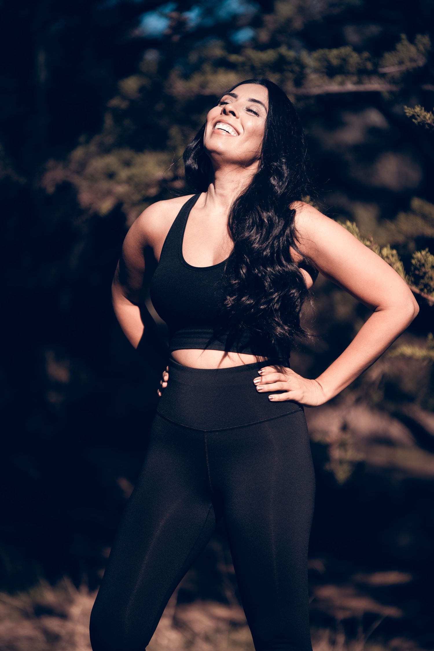 Athletic woman in black workout attire smiles while posing outdoors in natural lighting.