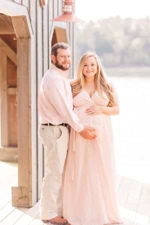 A maternity portrait of a couple in light pink attire on a wooden deck by the water.