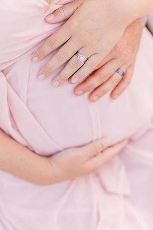 Close-up of engagement ring and hands resting on pink maternity dress.