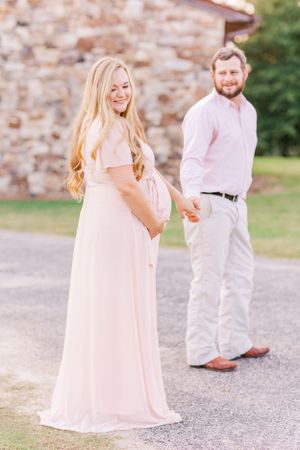 Expectant couple in coordinating pink and white outfits pose by stone wall.