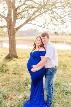 A maternity photoshoot in a field showing a couple embracing in front of a tree with the woman wearing a royal blue gown.