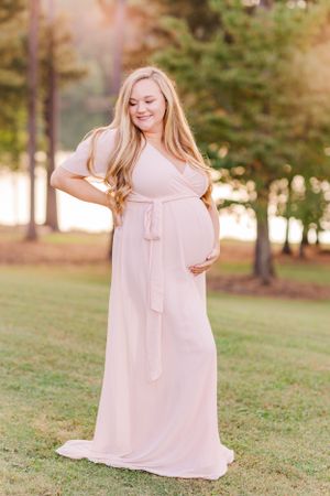 Expectant mother in flowing pink maxi dress poses in outdoor garden setting.