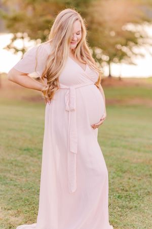 Pregnant woman in soft pink dress smiles while posing in sunlit outdoor setting.