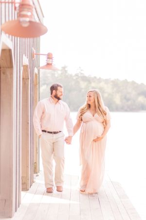 A sunny maternity photo of a couple walking on a waterfront deck in matching pink outfits.