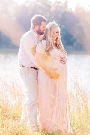 A dreamy maternity photo in golden light with the couple standing in tall grass wearing pink.