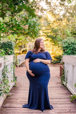 Maternity portrait on wooden bridge with woman in navy blue dress surrounded by garden scenery.