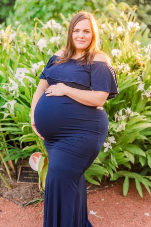 Maternity portrait in garden setting with woman wearing navy off-shoulder gown among flowering plants.