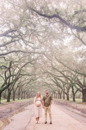 A couple walking down a dreamy path lined with overhanging oak trees in a romantic maternity photoshoot setting.